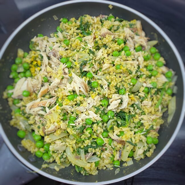 This image is showing a cooking pan containing a freshly cooked meal of flaked mackerel, brown rich, green peas, spinach and onion all mixed up to make mackerel kedgeree.